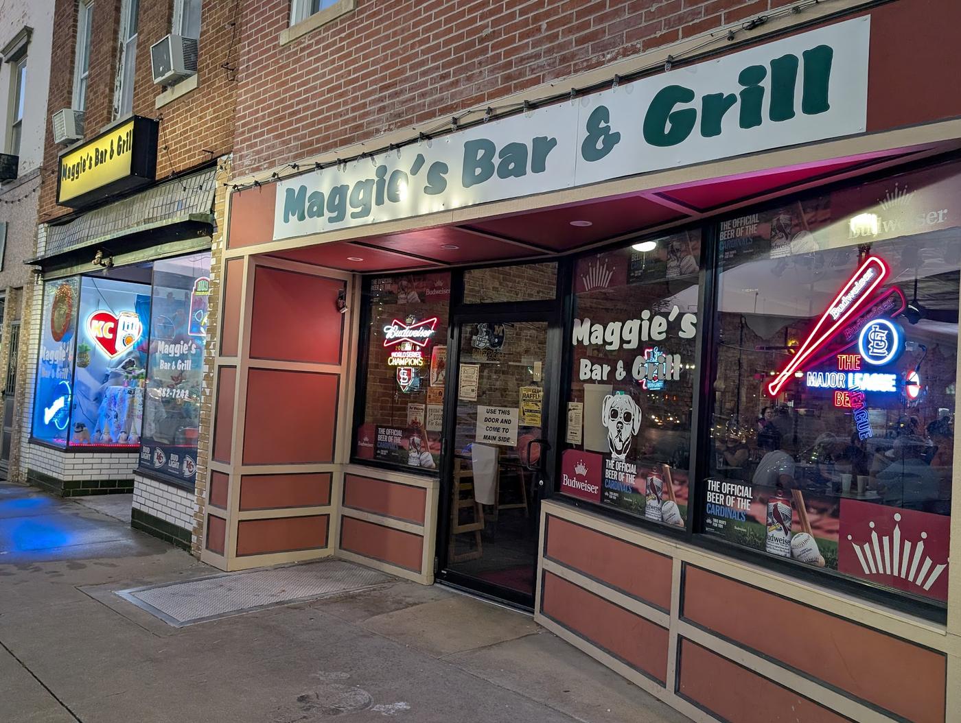 The Maggie's Bar & Grill storefront on Main Street at dusk: the name in green script on a white sign, brick, and a front window full of beer neon.