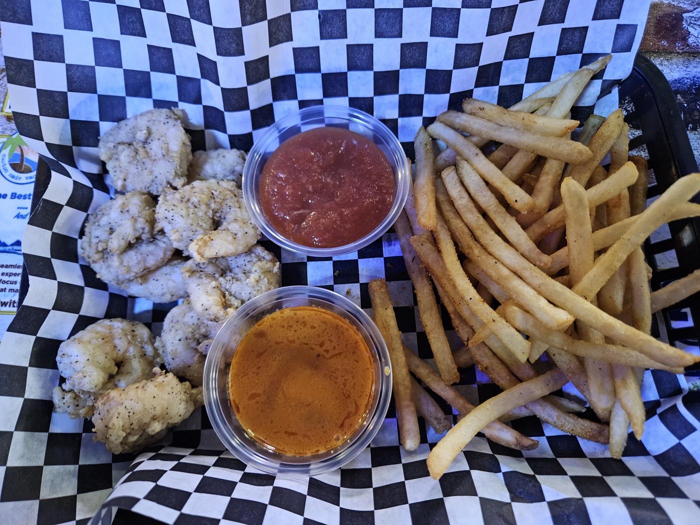 A basket of breaded, fried pieces with french fries and two dipping sauces on black and white checkered paper.