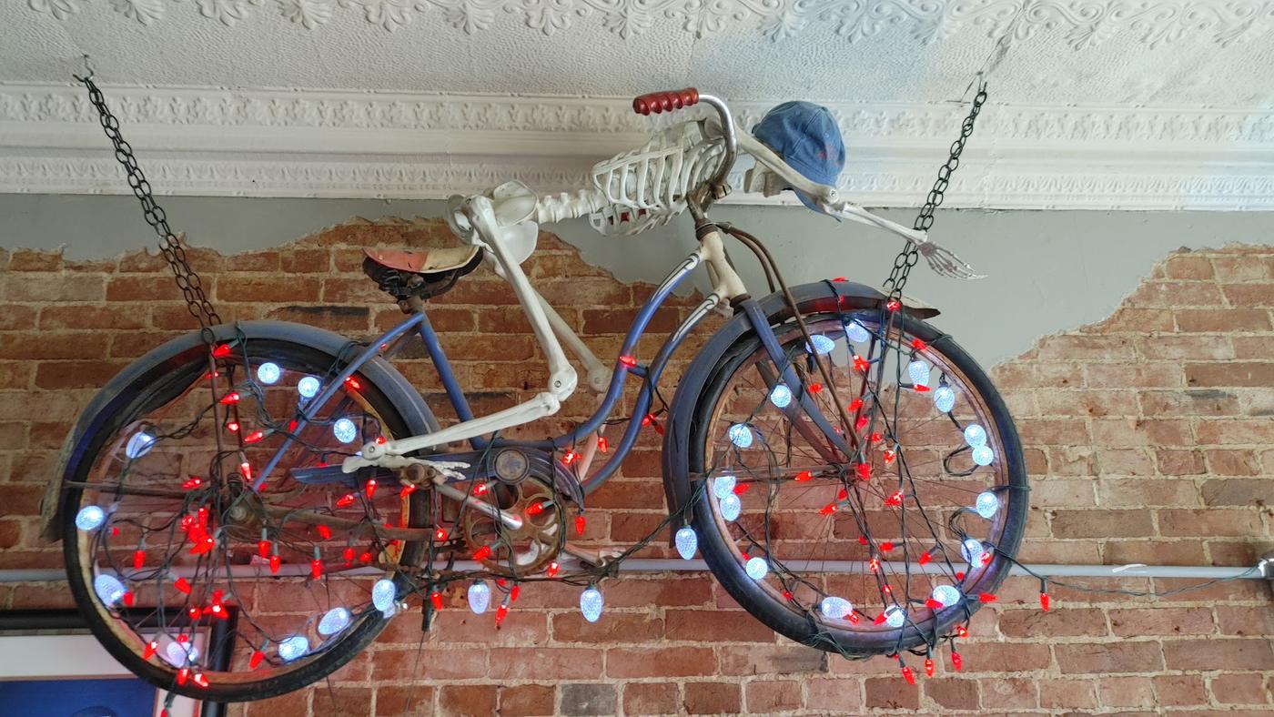 A vintage bicycle hung from the ceiling with a skeleton riding it, strung with red and white lights against exposed brick.